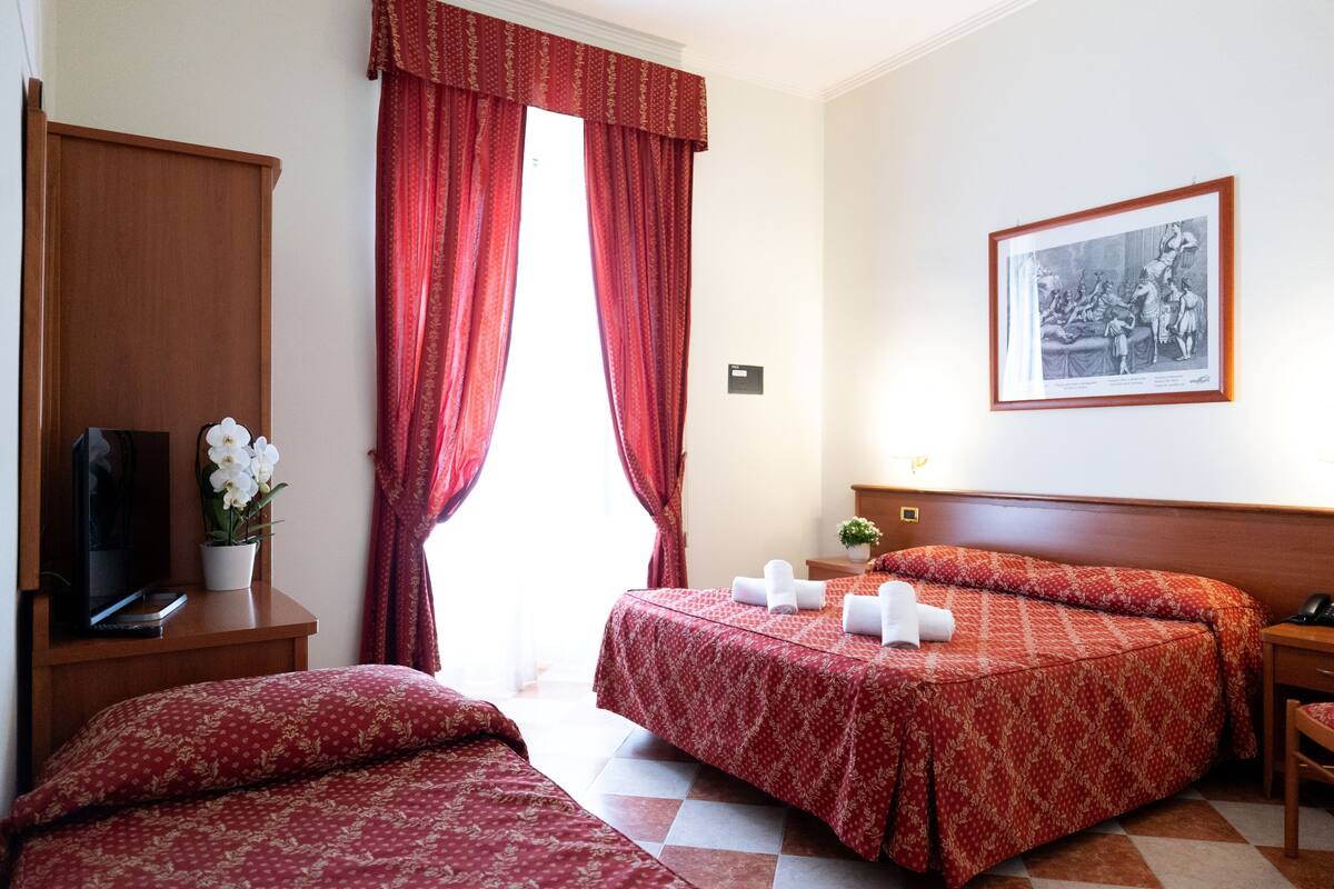 A hotel room with two beds, a television, a plant, and a framed picture on the wall.