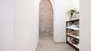 A narrow hallway with a brick wall, a white door, and a shelf with white dishes and a vase of flowers. - Hotel RomAntica (Rome)