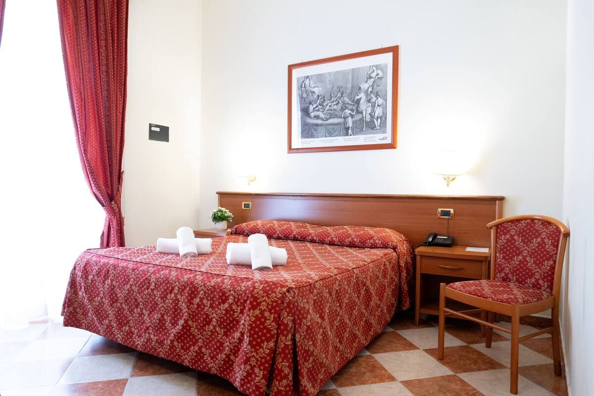 A hotel room with a bed, a desk, a chair, a framed picture on the wall, and a window with curtains.