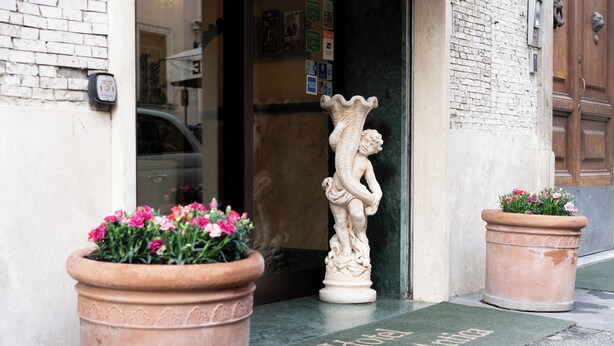 A hotel entrance with a doormat, terracotta planters with flowers, and a stone statue. - Hotel RomAntica (Rome)