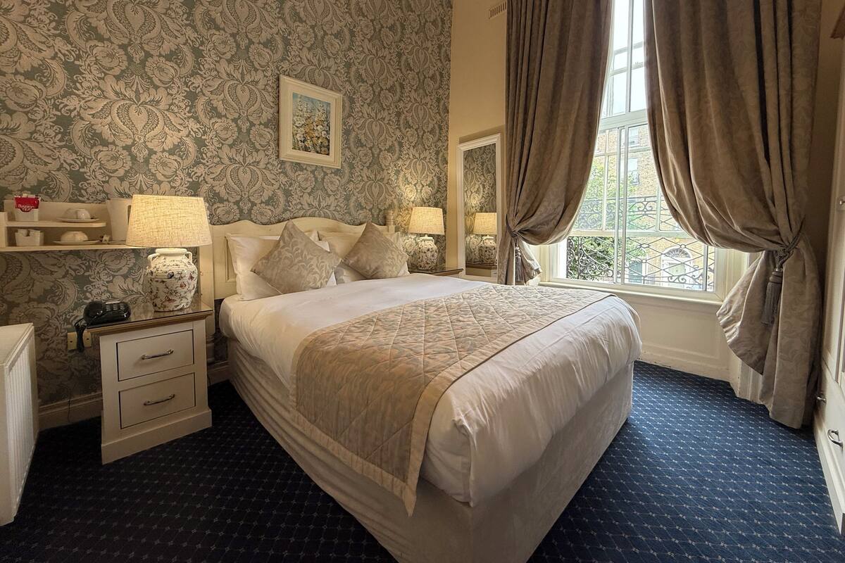 A neatly arranged bedroom with a large bed, bedside tables, a lamp, and a window with curtains.