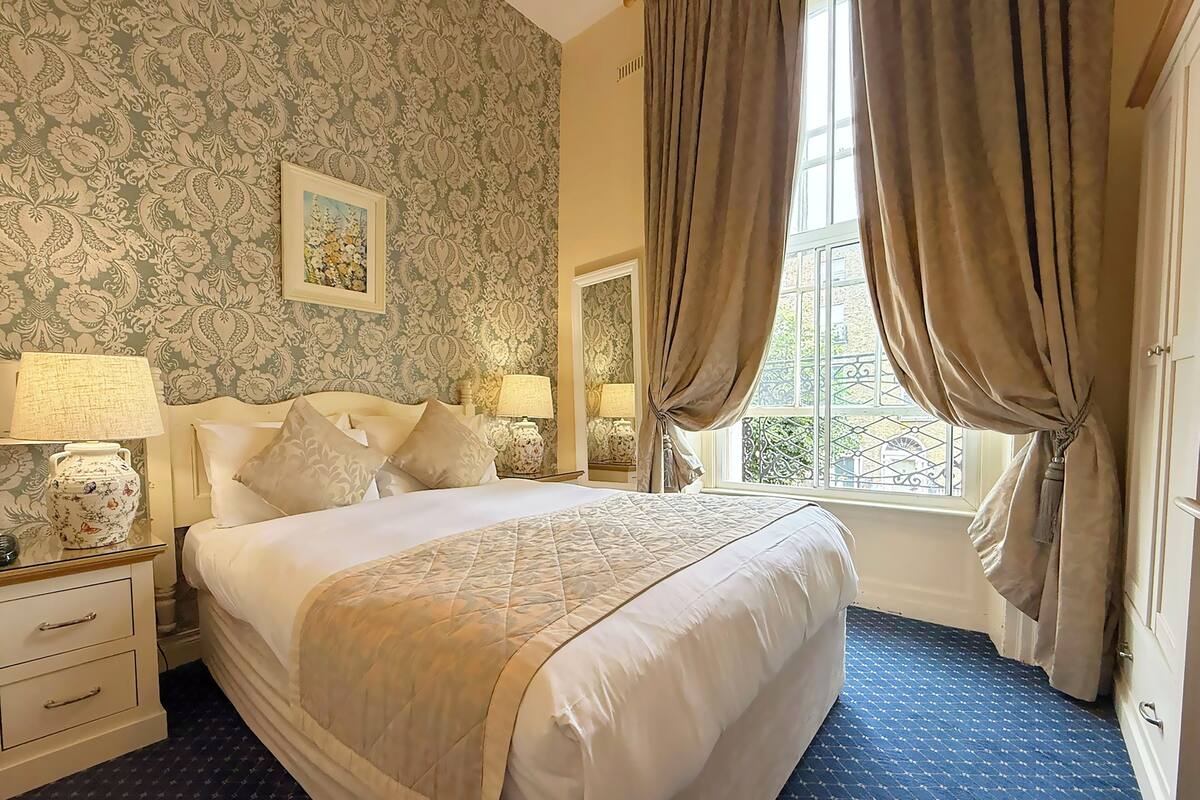 A neatly made bed with a quilt, two bedside tables with lamps, a large window with curtains, and a patterned wallpaper.