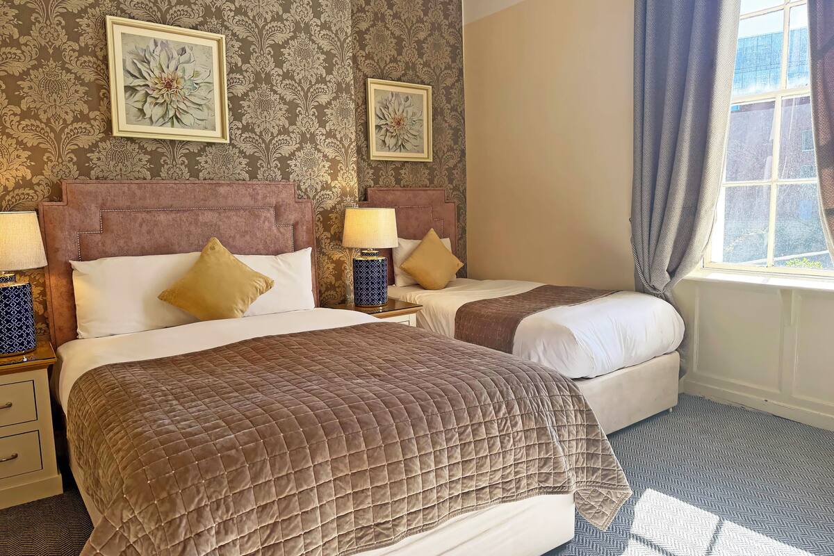 A hotel room with two beds, a nightstand with a lamp, a dresser, and a window with curtains.