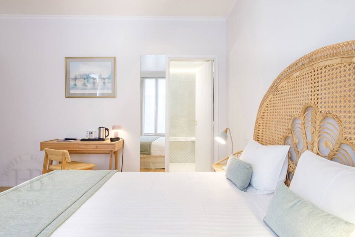 A hotel room with a bed, a desk, a chair, a lamp, and a door leading to another room.