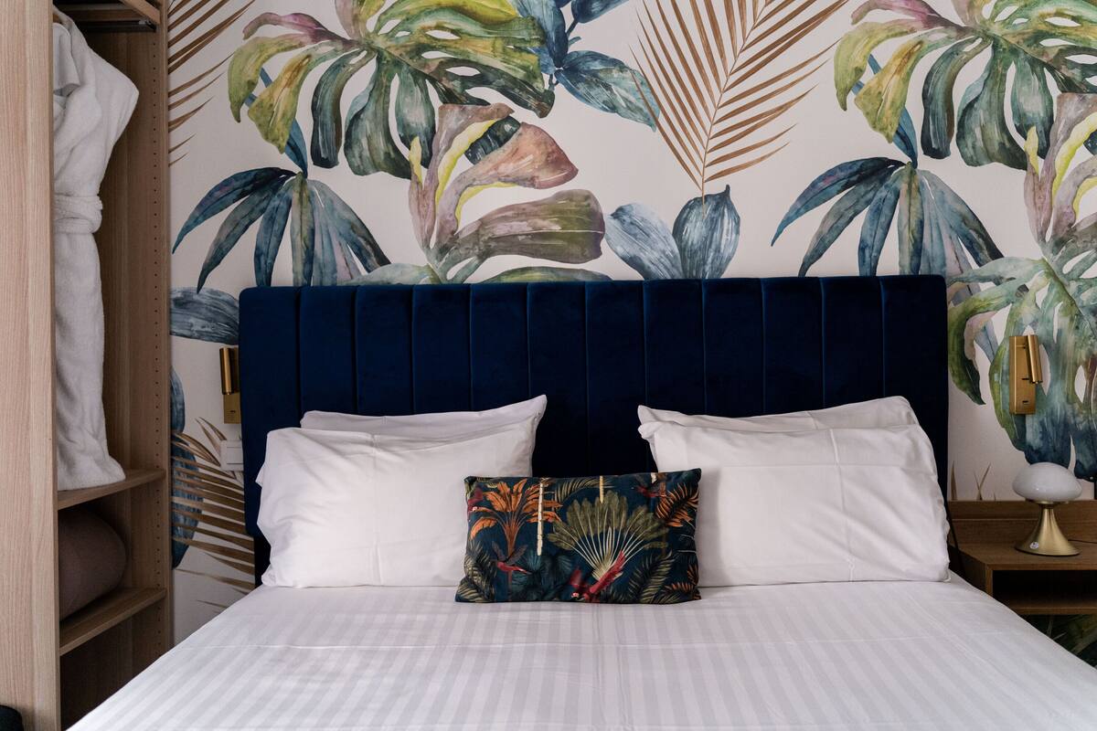 A bed with white linens, a dark blue headboard, and a tropical leaf-patterned pillow.