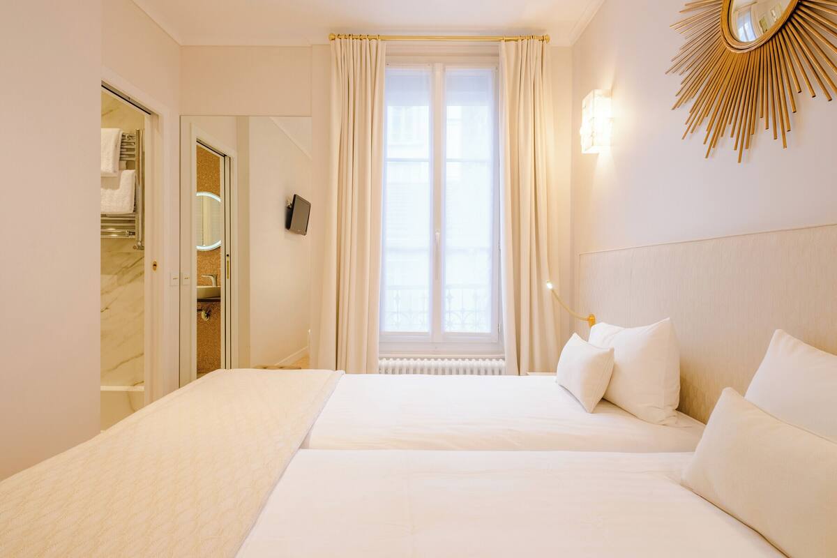 A hotel room with a large bed, a window with curtains, a mirror, and a wall-mounted light fixture.