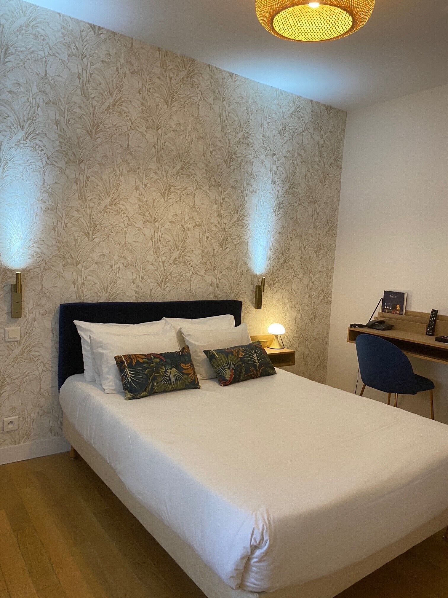 Una cama bien hecha con sábanas blancas y dos almohadas decorativas, un escritorio de madera con una silla y una lámpara fijada a la pared.