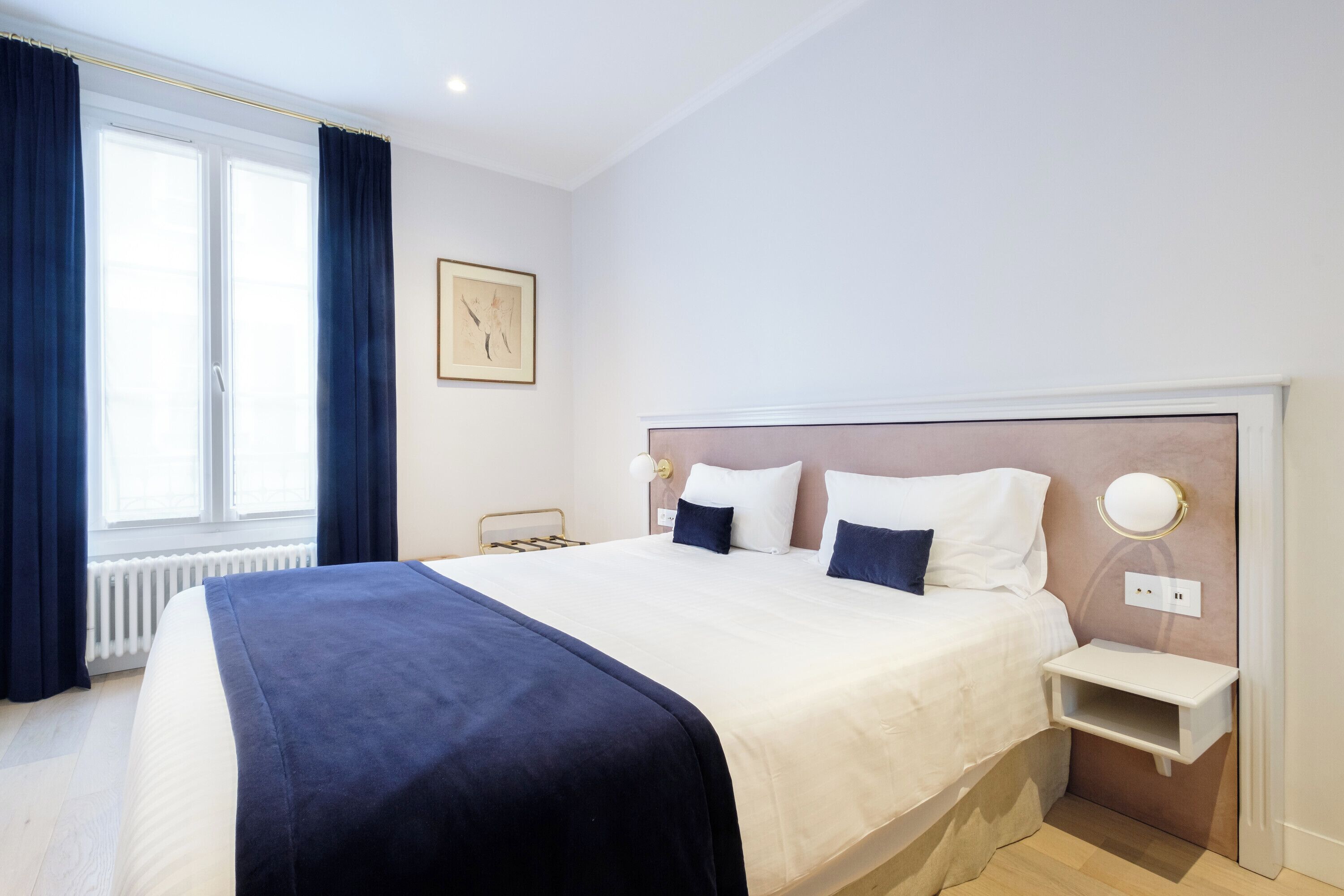 Quarto de hotel bem arrumado com uma cama grande, mesa de cabeceira, luminária na parede e uma janela com cortinas.