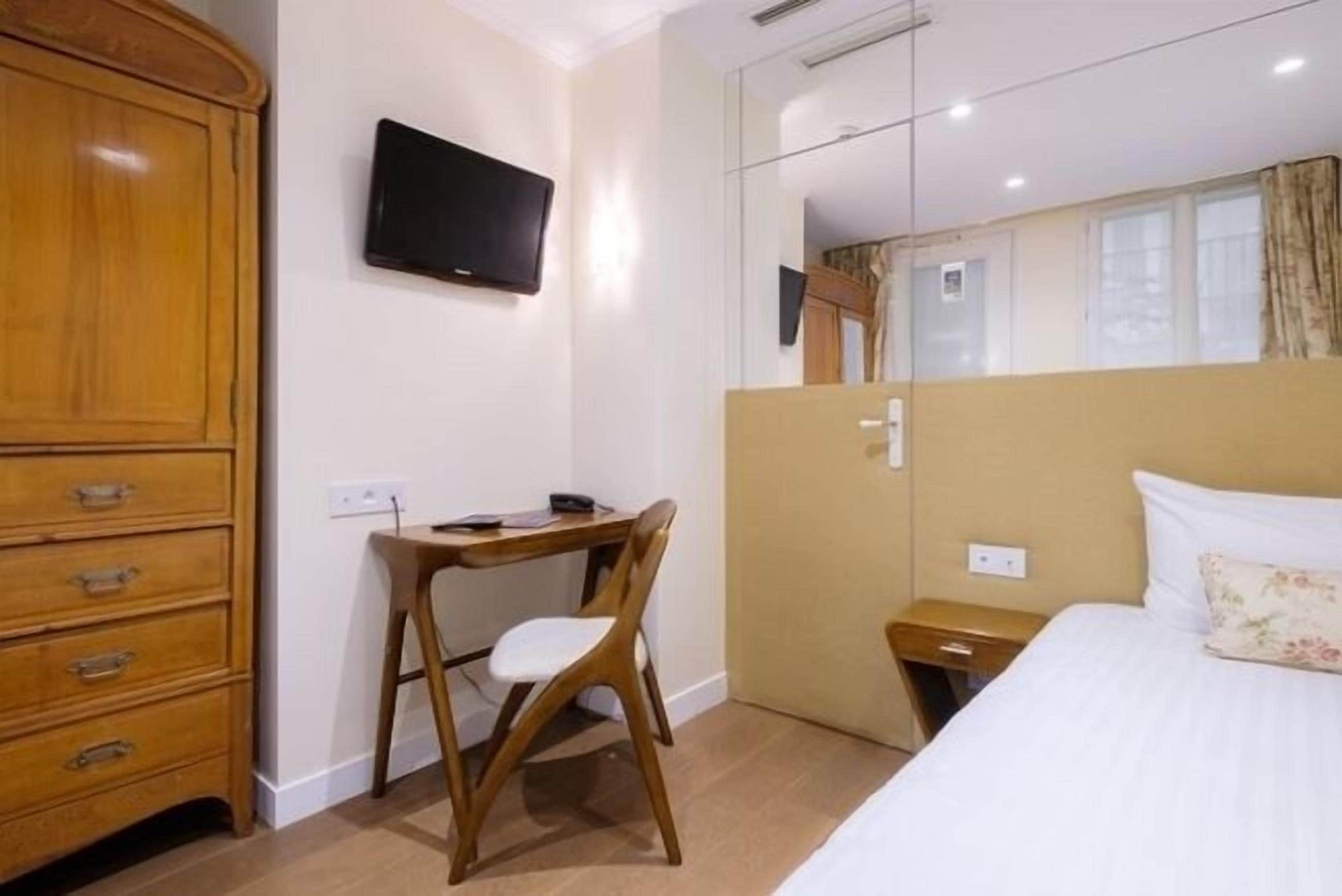 A hotel room with a wooden wardrobe, a desk with a chair, a bed with a pillow, a TV mounted on the wall, and a mirror reflecting part of the room.