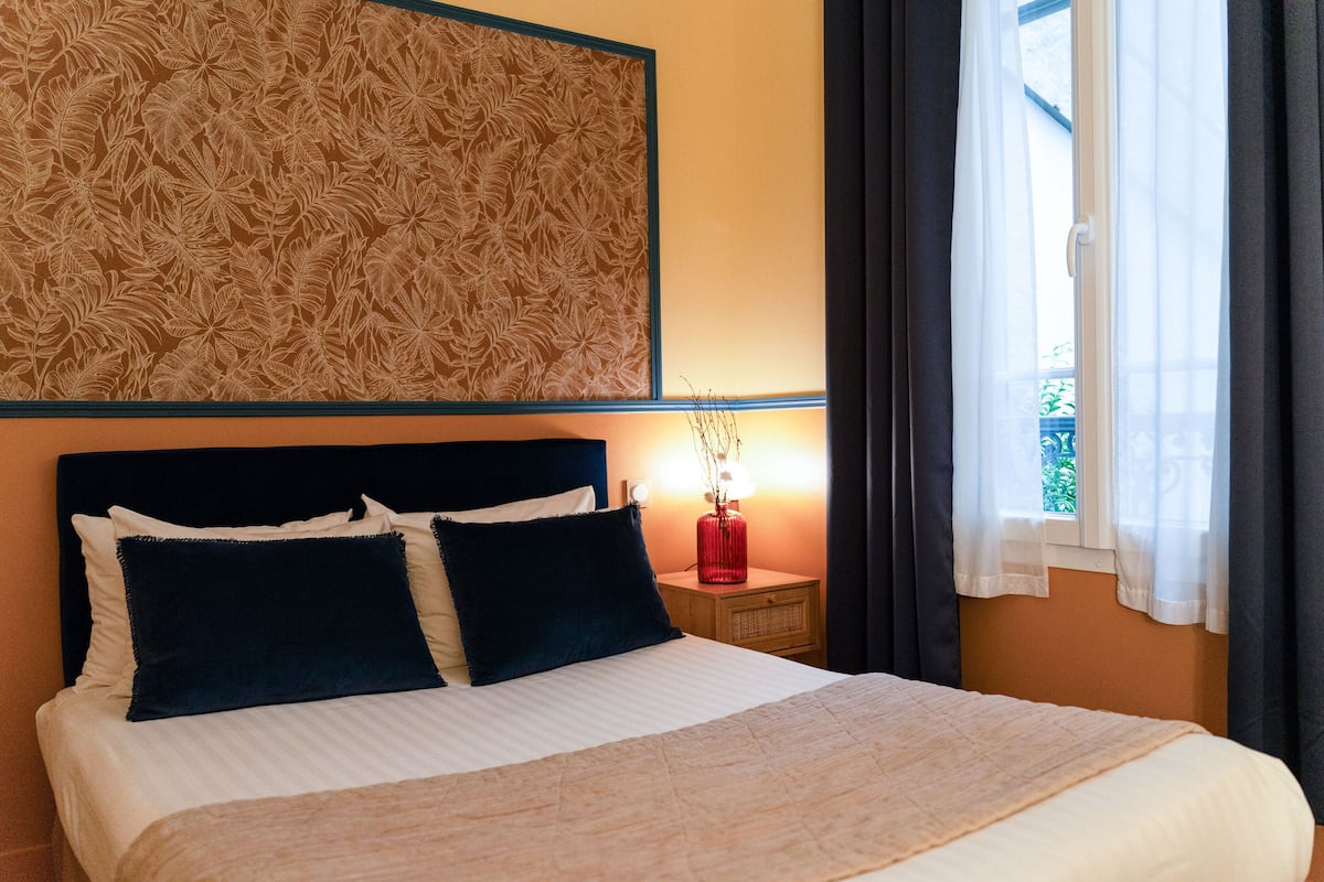 A neatly made bed with a white and beige duvet, dark blue pillows, and a headboard with a leaf pattern. A small red lamp is on a bedside table next to the bed.
