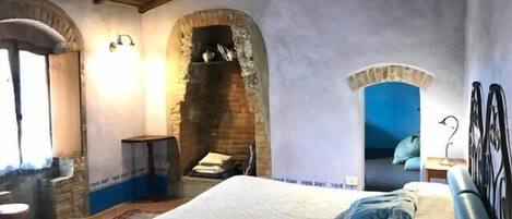 A bedroom with a stone fireplace, a bed with a blue pillow, and a view of a blue sky through an arched window.
