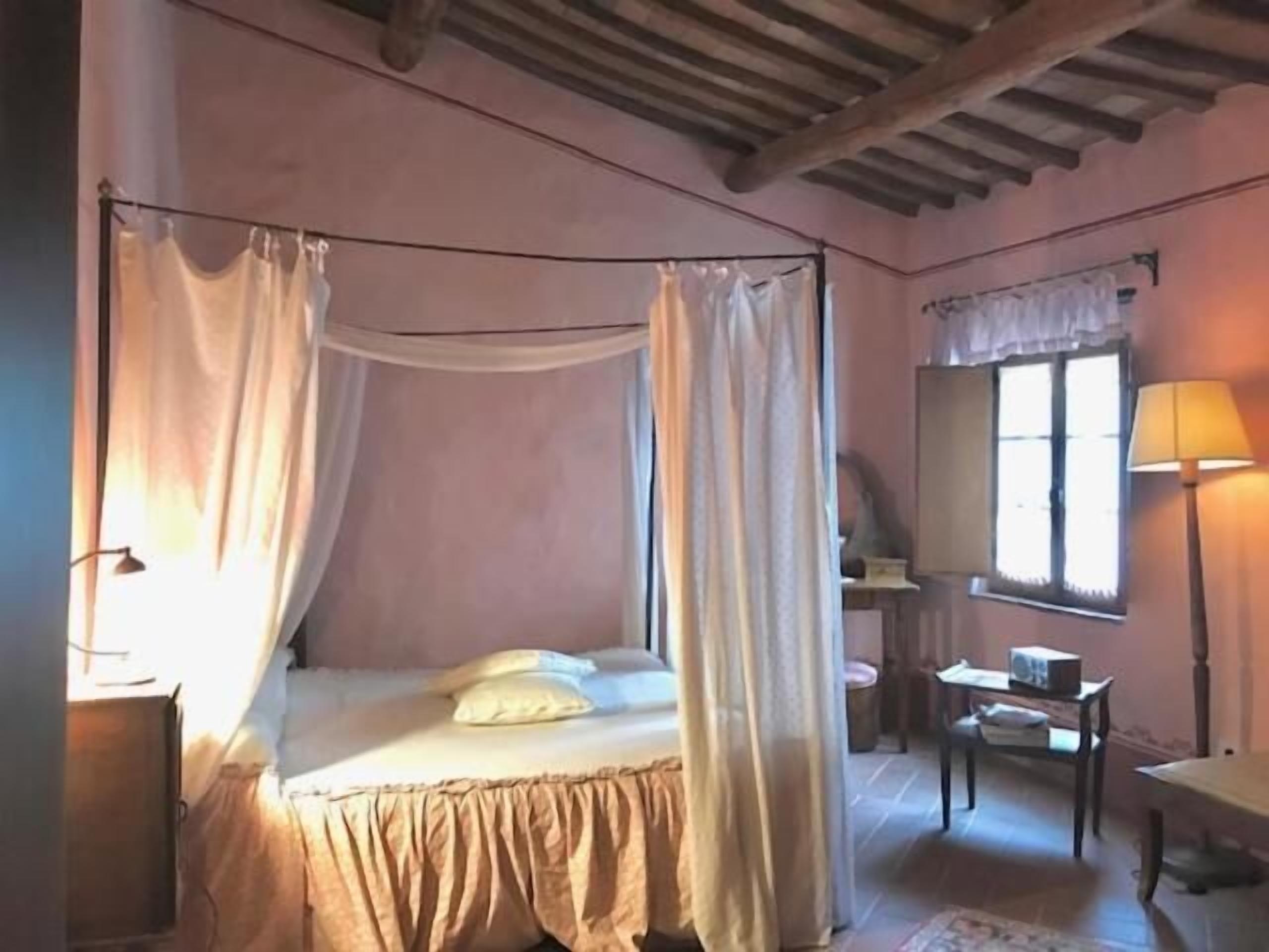 Un dormitorio con cama con dosel, una mesita de noche, una lámpara y una ventana con cortinas.