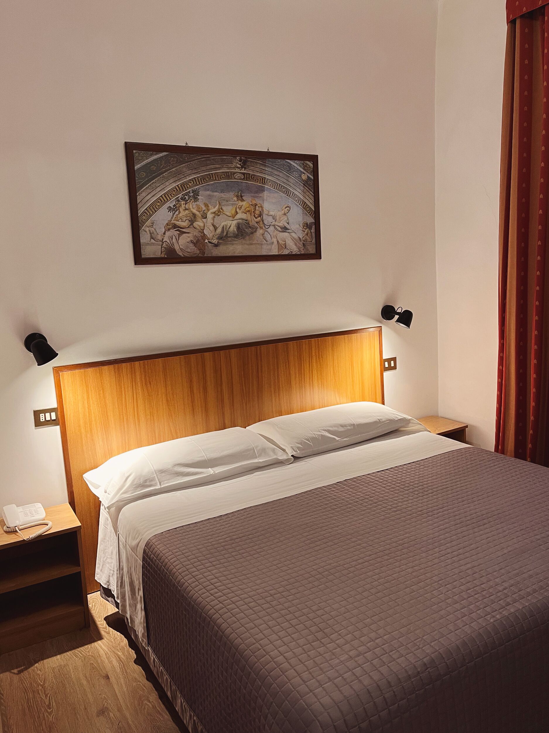 A hotel room with a wooden headboard, a bed with white linens, a nightstand with a phone, and a painting above the bed.