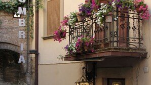 Entrada de un hotel con un balcón decorado con flores, un letrero que indica el nombre del hotel y una pared de piedra a la izquierda.