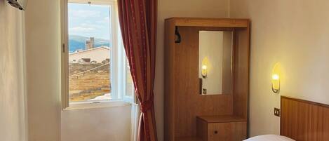 Habitación de hotel con cama, mesita de noche de madera, espejo, ventana con cortinas y vistas al exterior.