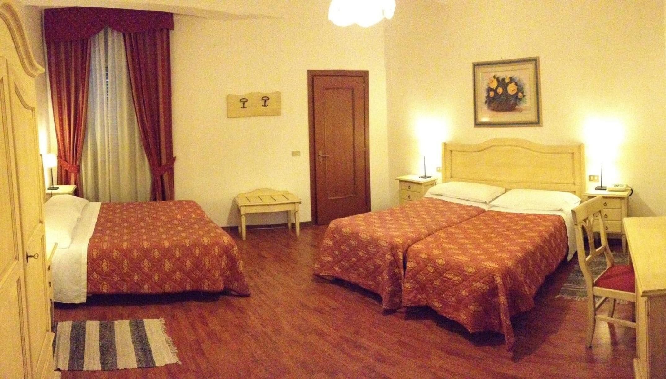 A hotel room with two beds, a wooden floor, a small table, a chair, a door, and a wall-mounted light fixture.