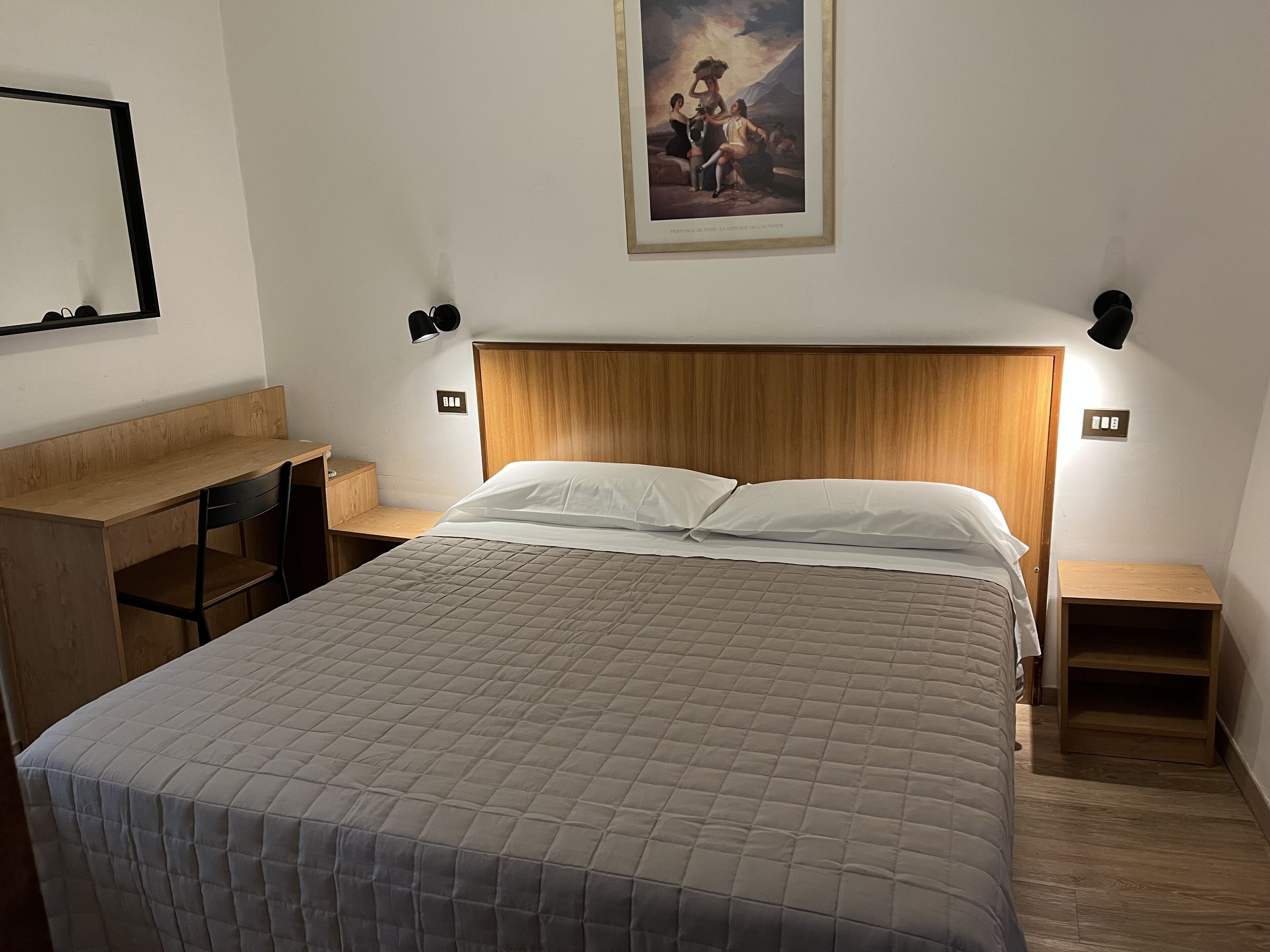 A hotel room with a wooden headboard, a bed with a quilted cover, a desk with a chair, and a small bedside table.