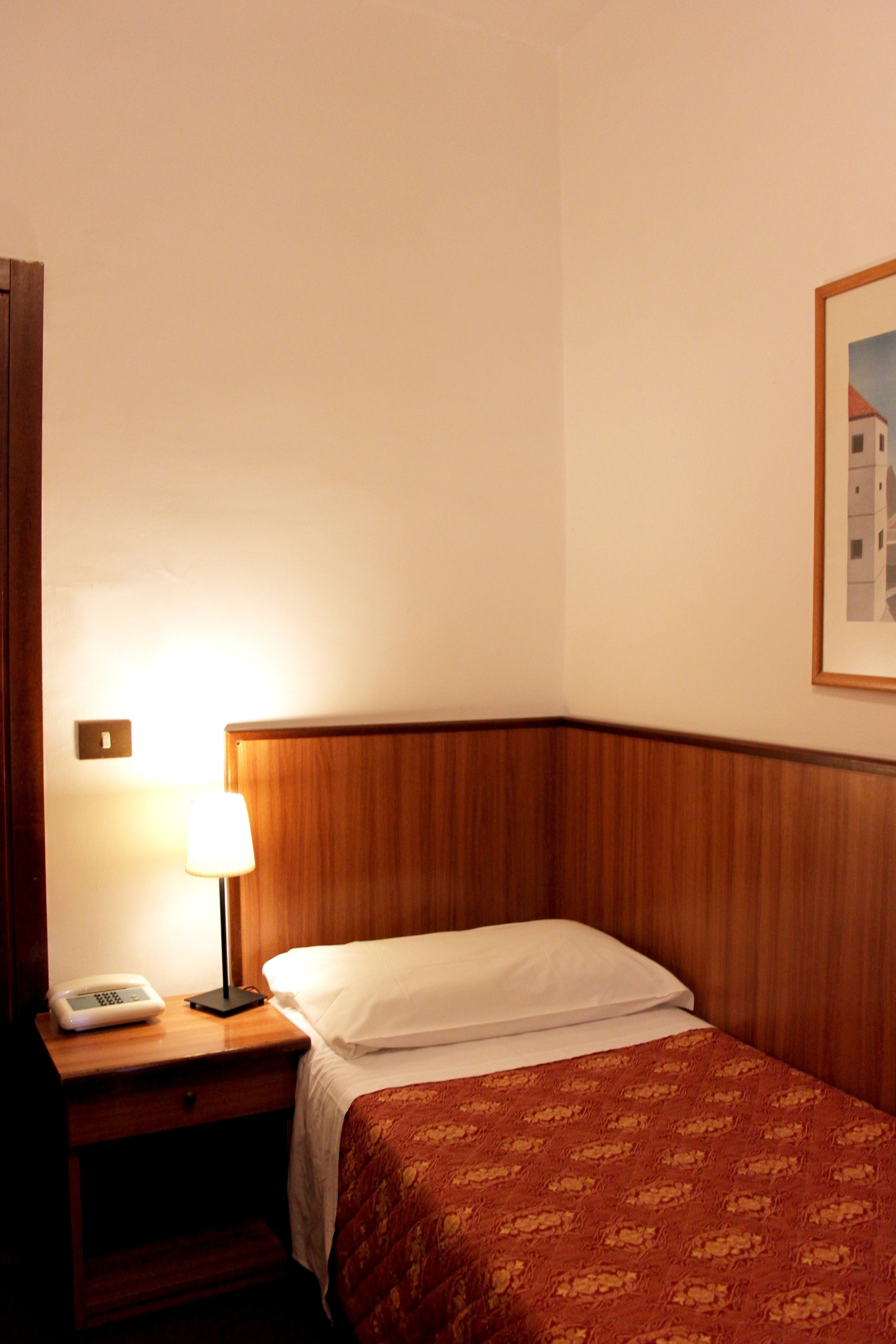 A hotel room with a wooden headboard, a bedside table with a lamp, a phone, and a bed with a patterned blanket.