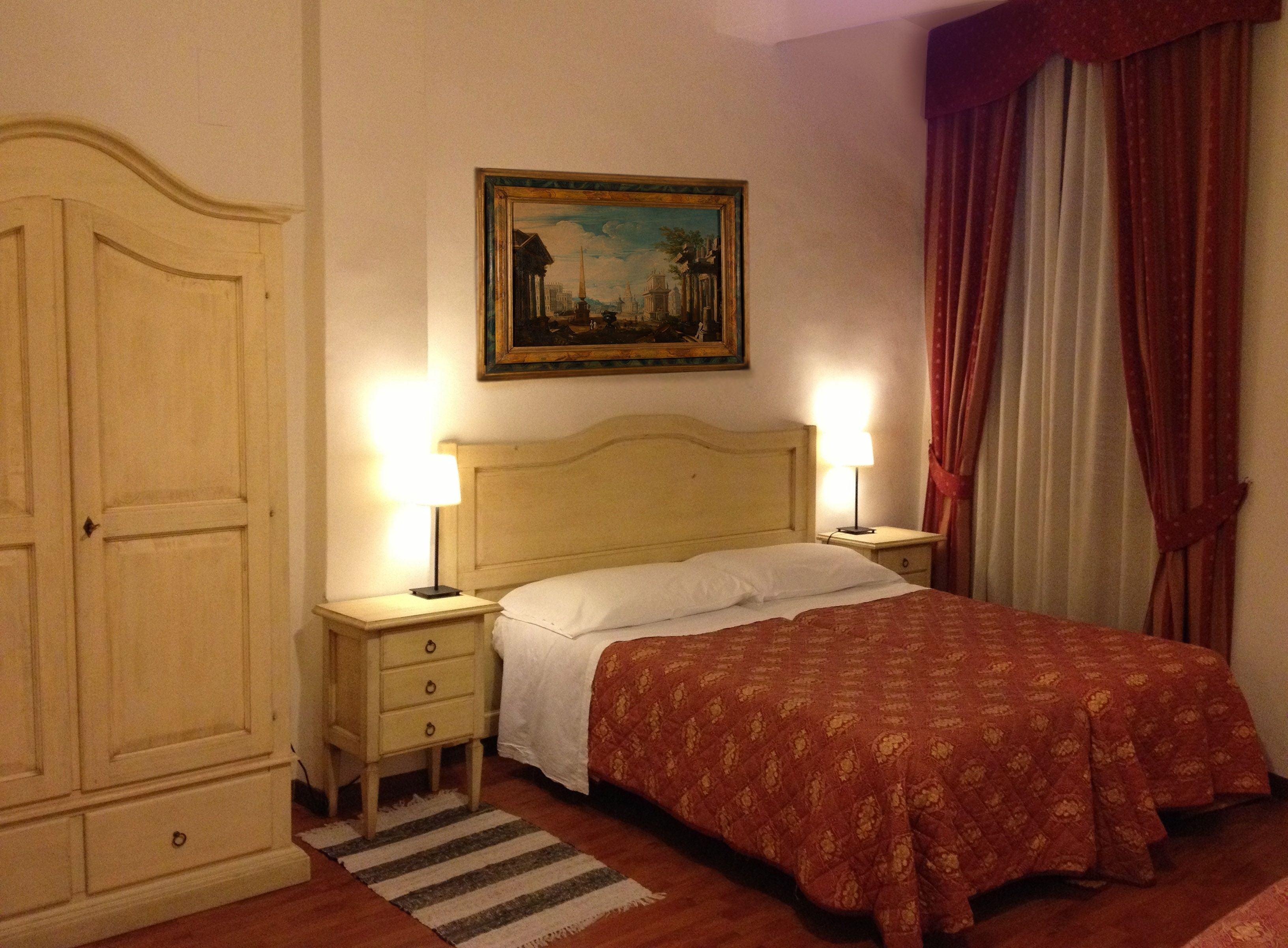 A hotel room with a bed, two bedside tables with lamps, a wardrobe, a painting on the wall, and a window with curtains.