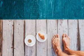 A wooden deck with a pair of feet, a cup of coffee, and a croissant.