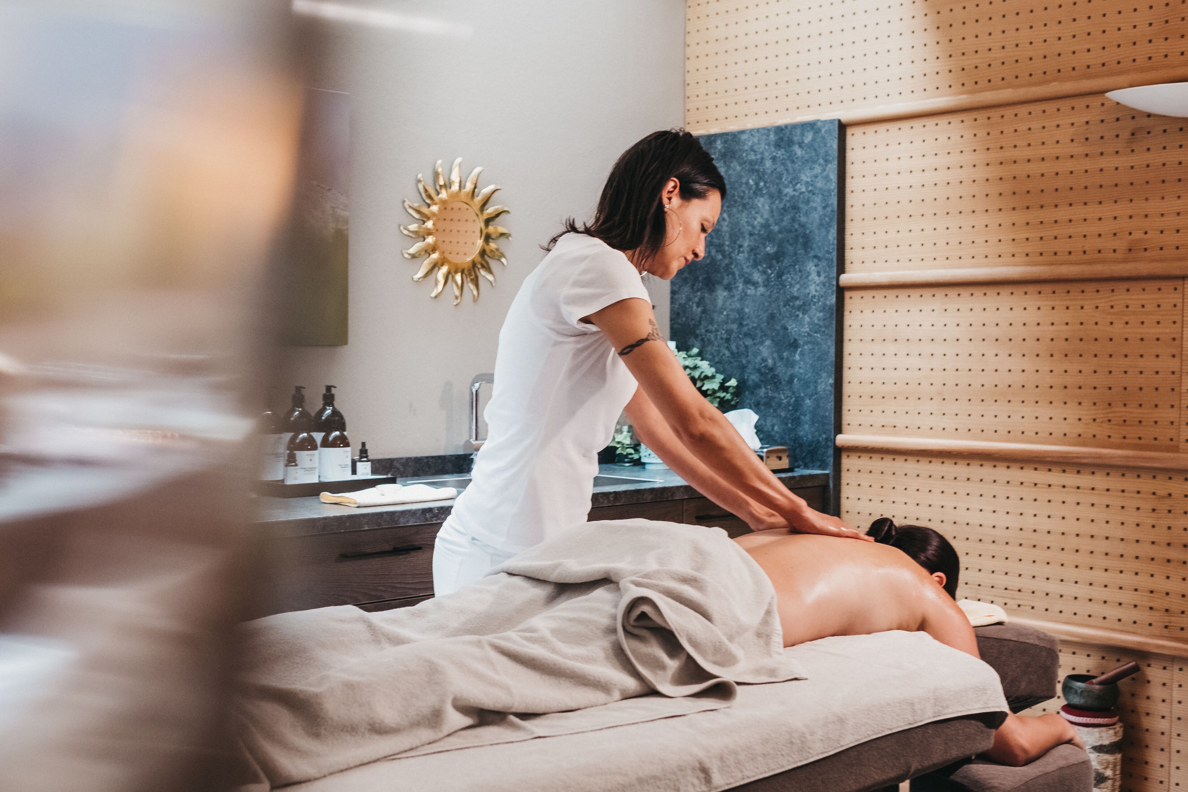 A massage therapy room with a massage table, a client receiving a back massage, and a wall-mounted shelf with bottles and a decorative sunburst mirror.