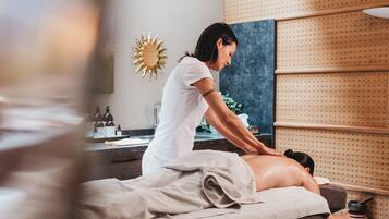 A massage therapy room with a massage table, a client receiving a back massage, and a wall-mounted shelf with bottles and a decorative sunburst mirror.
