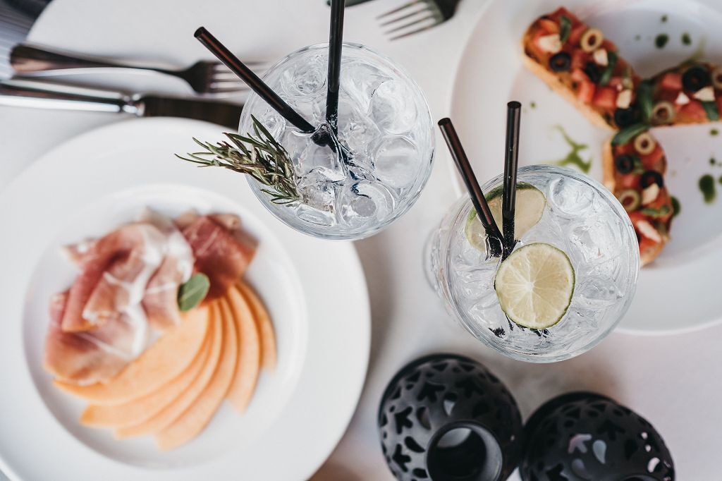 A table setting with two glasses of a clear beverage with ice and garnishes, a plate of sliced melon, and a pizza with toppings.