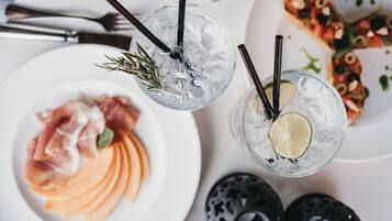 A table setting with two glasses of a clear beverage with ice and garnishes, a plate of sliced melon, and a pizza with toppings.