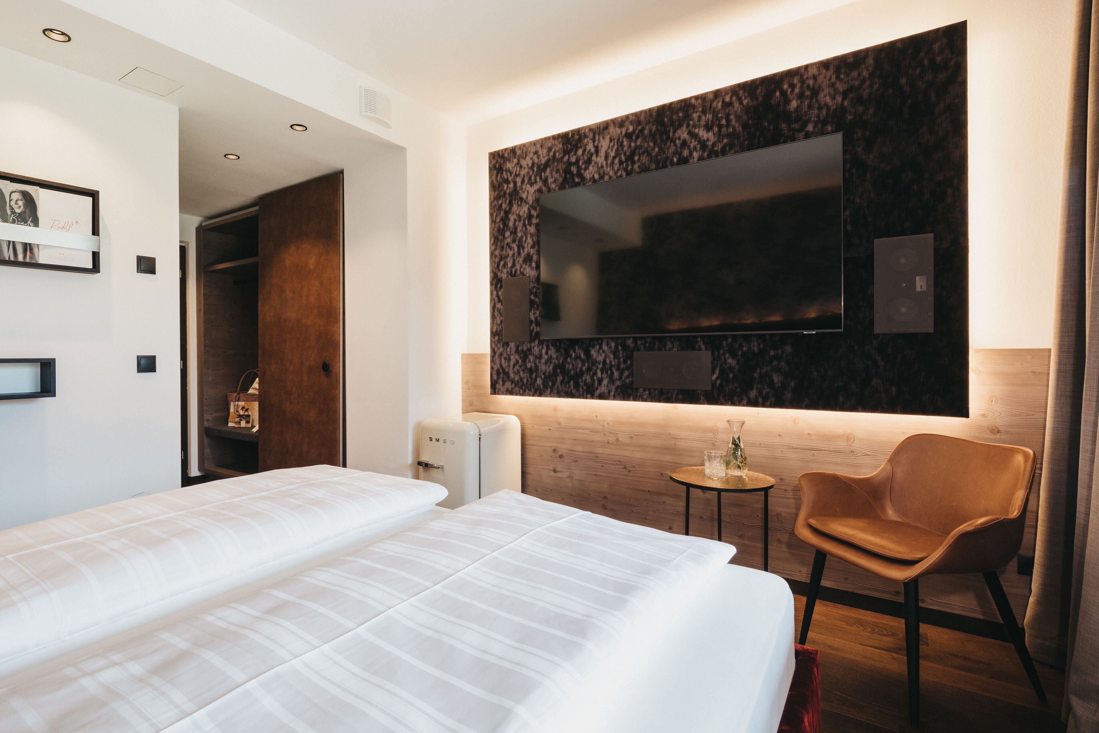 A hotel room with a large bed, a TV mounted on the wall, a small table with a chair, and a wooden wardrobe.