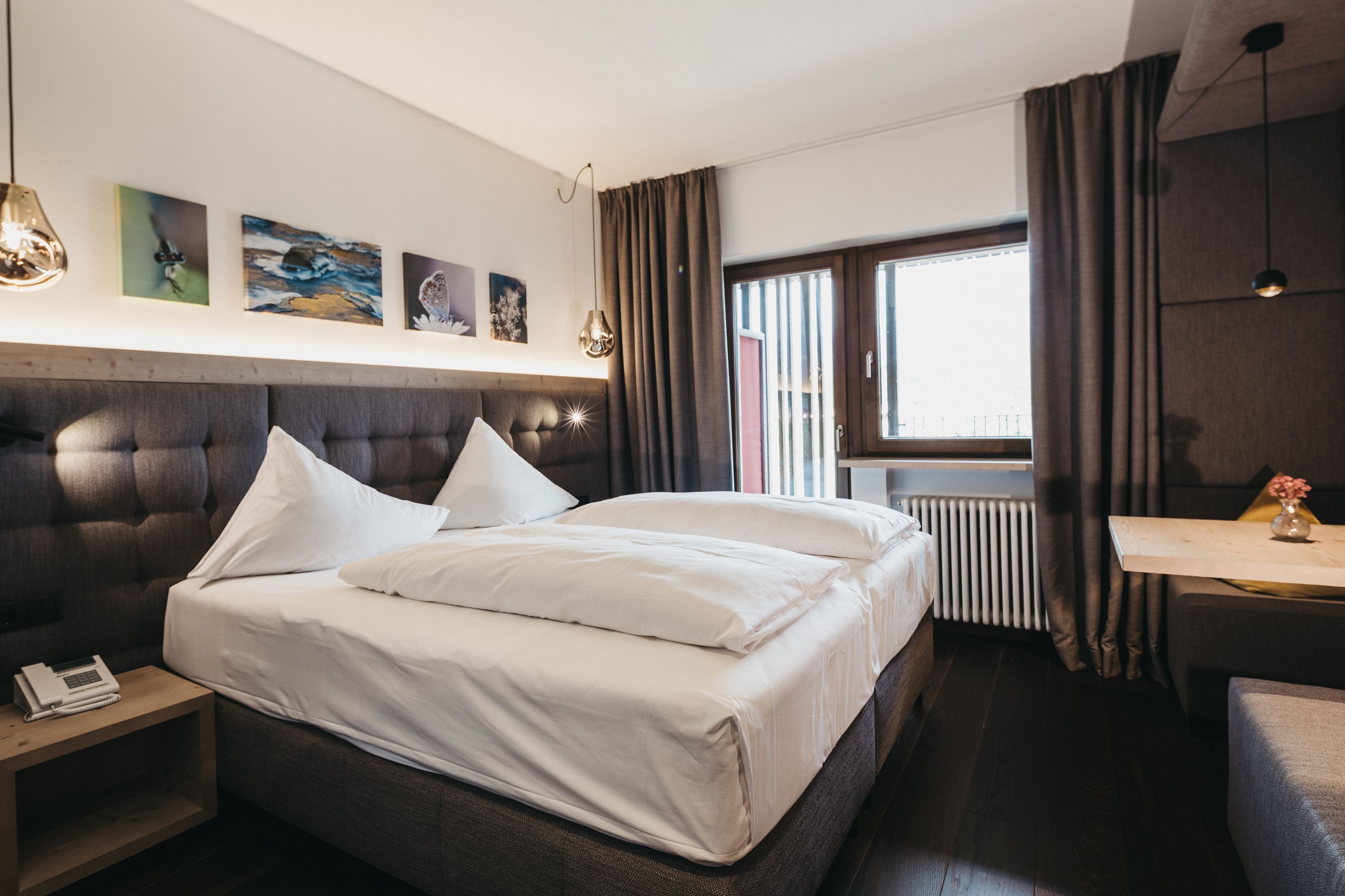 Ein modernes Hotelzimmer mit einem großen Bett, einem Nachttisch mit Telefon, einem kleinen Schreibtisch mit Vase und einem Fenster mit Vorhängen.