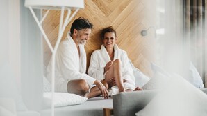 A couple sitting on a bed in a modern room with a wooden wall and a white ceiling. - Hotel Rudolf (Brunico)