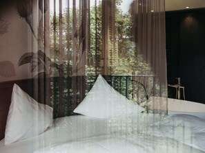 A bedroom with a bed, a window with sheer curtains, and a view of trees and buildings outside.