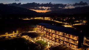 A modern hotel with multiple floors, outdoor seating, and a view of mountains in the background. - Hotel Rudolf (Brunico)