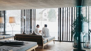 A modern hotel room with a large window, a sofa, a bedside table, and a lamp. - Hotel Rudolf (Brunico)