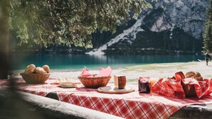 A picnic setup with bread, cheese, and juice on a checkered tablecloth by a lake with mountains in the background. - Hotel Rudolf (Brunico)