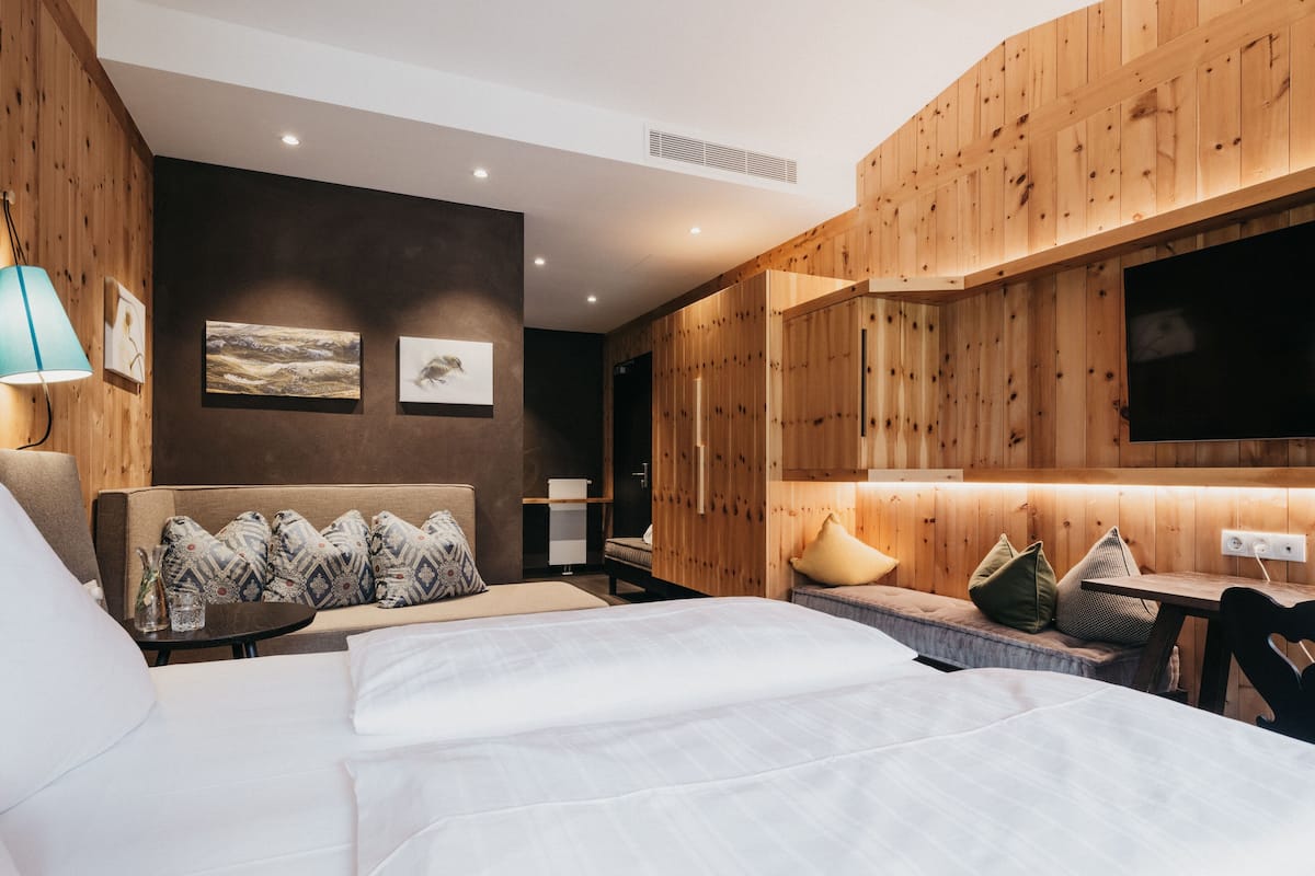 Ein modernes Hotelzimmer mit Wandpaneelen aus Holz, einem großen Bett, einer Sitzecke inklusive Sofa und Sesseln, einem am Wand montierten Flachbildfernseher und einem kleinen Tisch mit Lampe.