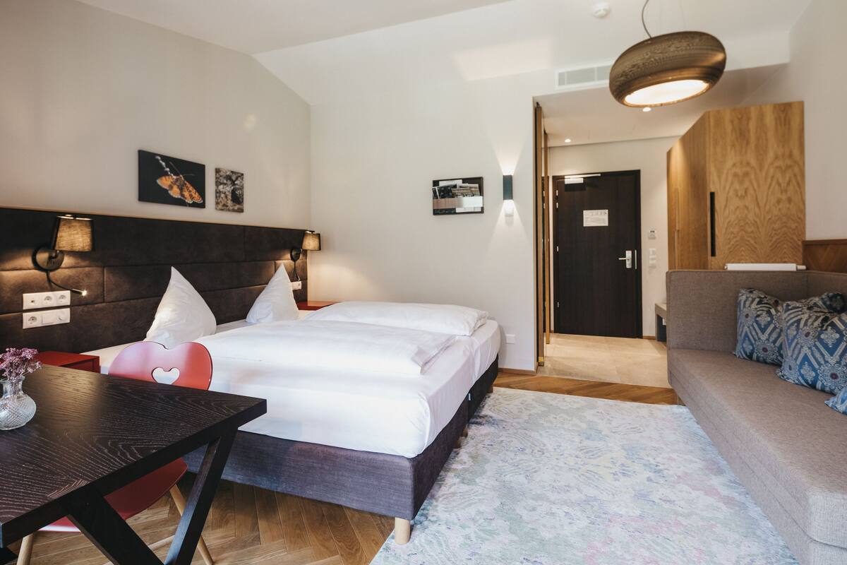 Ein modernes Hotelzimmer mit einem großen Bett, einem Schreibtisch, einem roten Sessel und einer Couch. An den Wänden sind Lampen angebracht und eine Holzvertäfelung sorgt für Akzente.