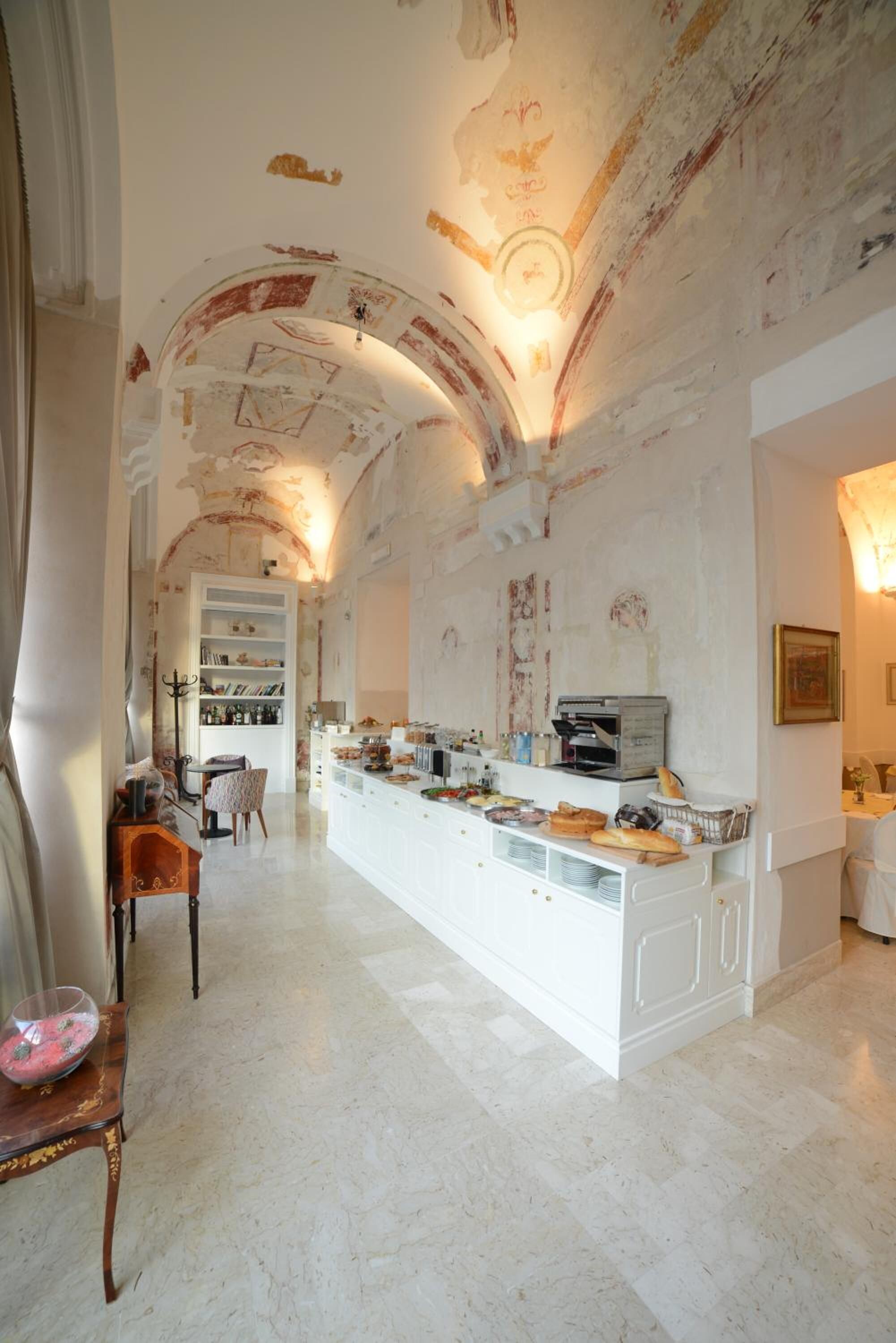 A spacious room with a high ceiling, a central island with food and drink, and a seating area with a chair and a table.