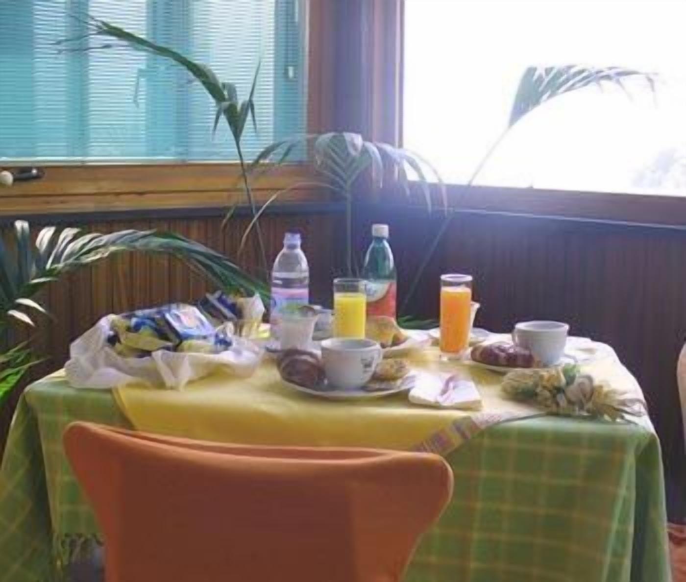 Une table de petit-déjeuner avec divers aliments et boissons, une chaise et une fenêtre avec des stores et des plantes.