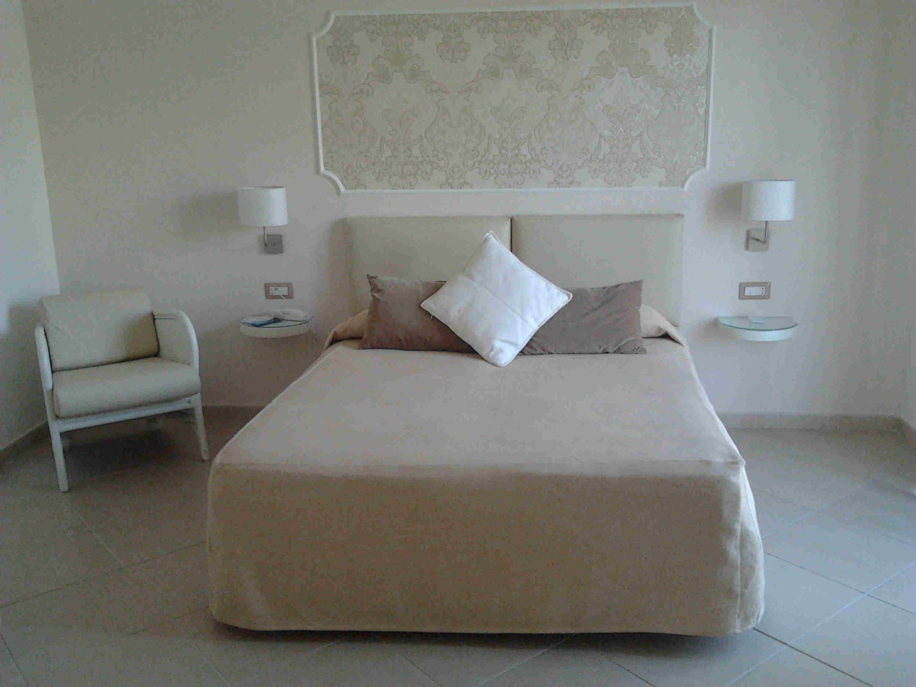 A bedroom with a bed, a chair, two bedside tables, and a wall-mounted light fixture.