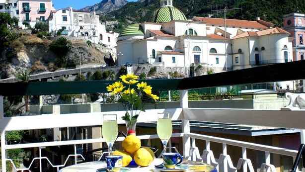 A balcony with a table set for two, featuring a vase with yellow flowers, a glass of drink, and a lemon. The background includes a mountainous landscape with white buildings and a green-domed structure. - Riviera B&B Rooms and Apartament SEA-VIEW (Maiori)