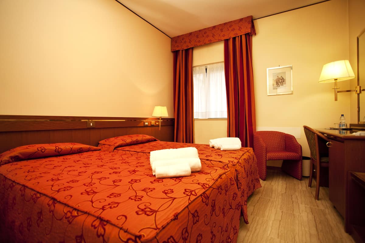 A hotel room with a large bed, a desk, a chair, a window with curtains, a lamp, and a framed picture on the wall.