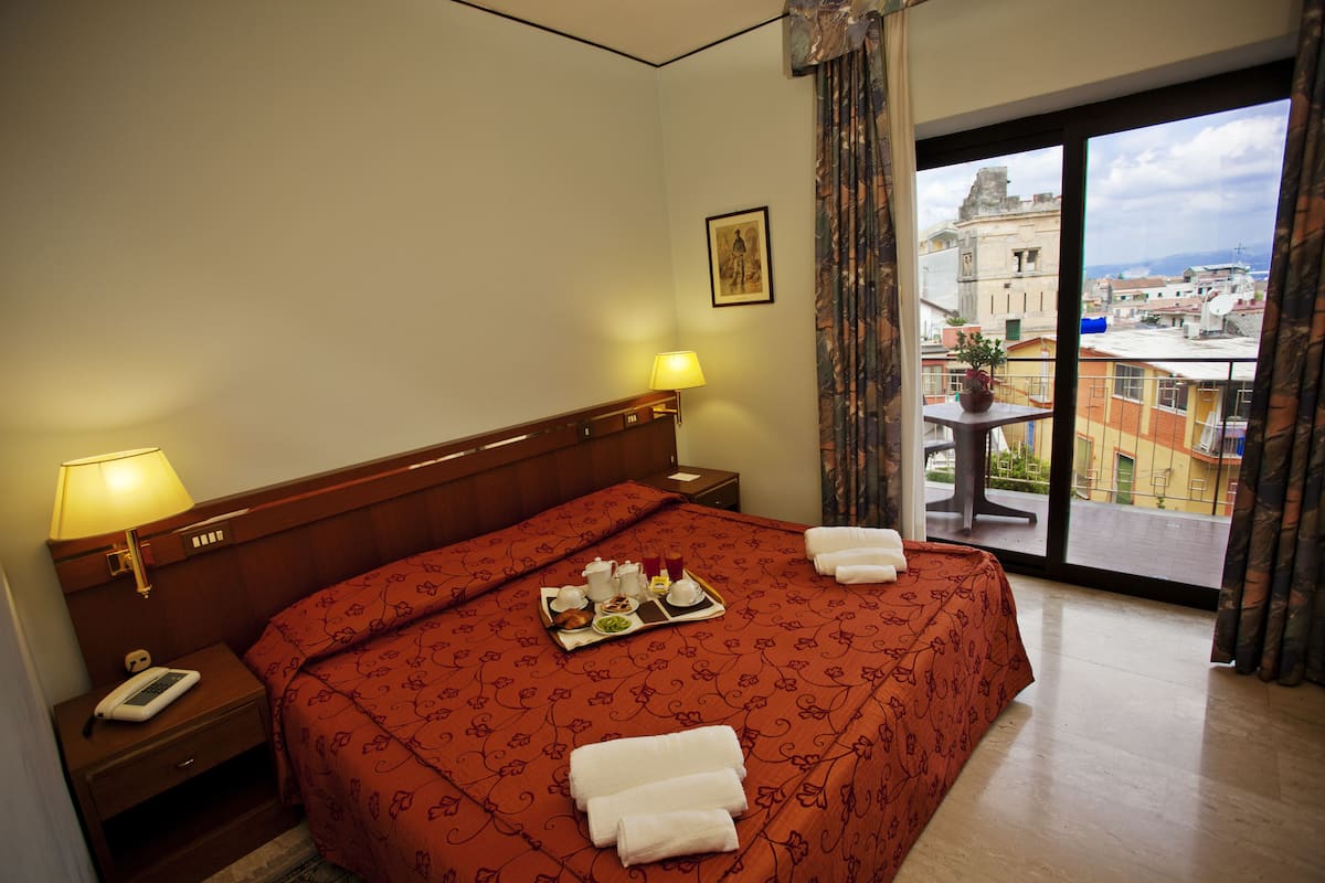 A hotel room with a large bed, two bedside lamps, a nightstand with a phone, a tray with breakfast items, and a balcony with a view of buildings and mountains.