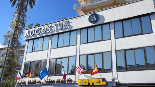 Ein Hotel namens "AUGUSTUS" mit mehreren Flaggen an der Außenfassade, darunter die EU-Flagge, die US-Flagge und weitere.