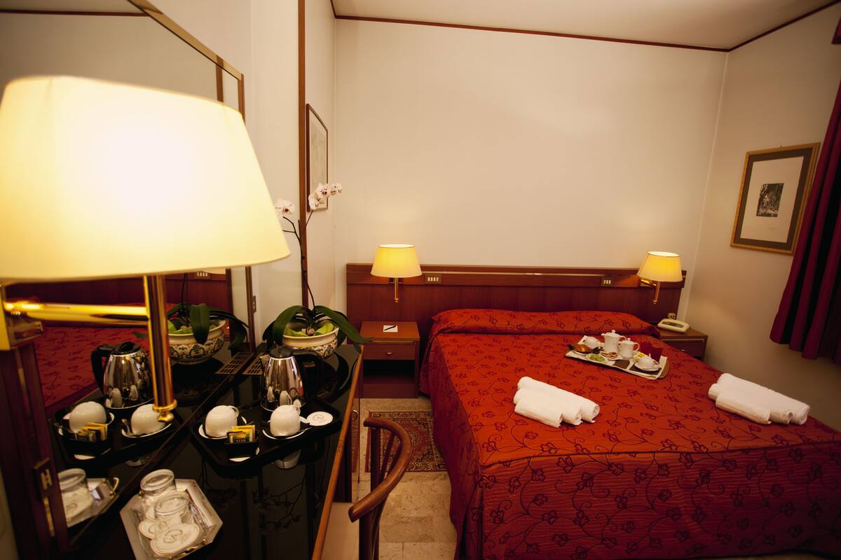 A hotel room with a bed, two bedside tables, a desk with a lamp, a chair, and a tray with breakfast items.