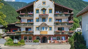 A hotel with a traditional Alpine architectural style, featuring multiple balconies with flower boxes, a central entrance, and a view of a mountainous landscape. - Gissbach (Brunico)