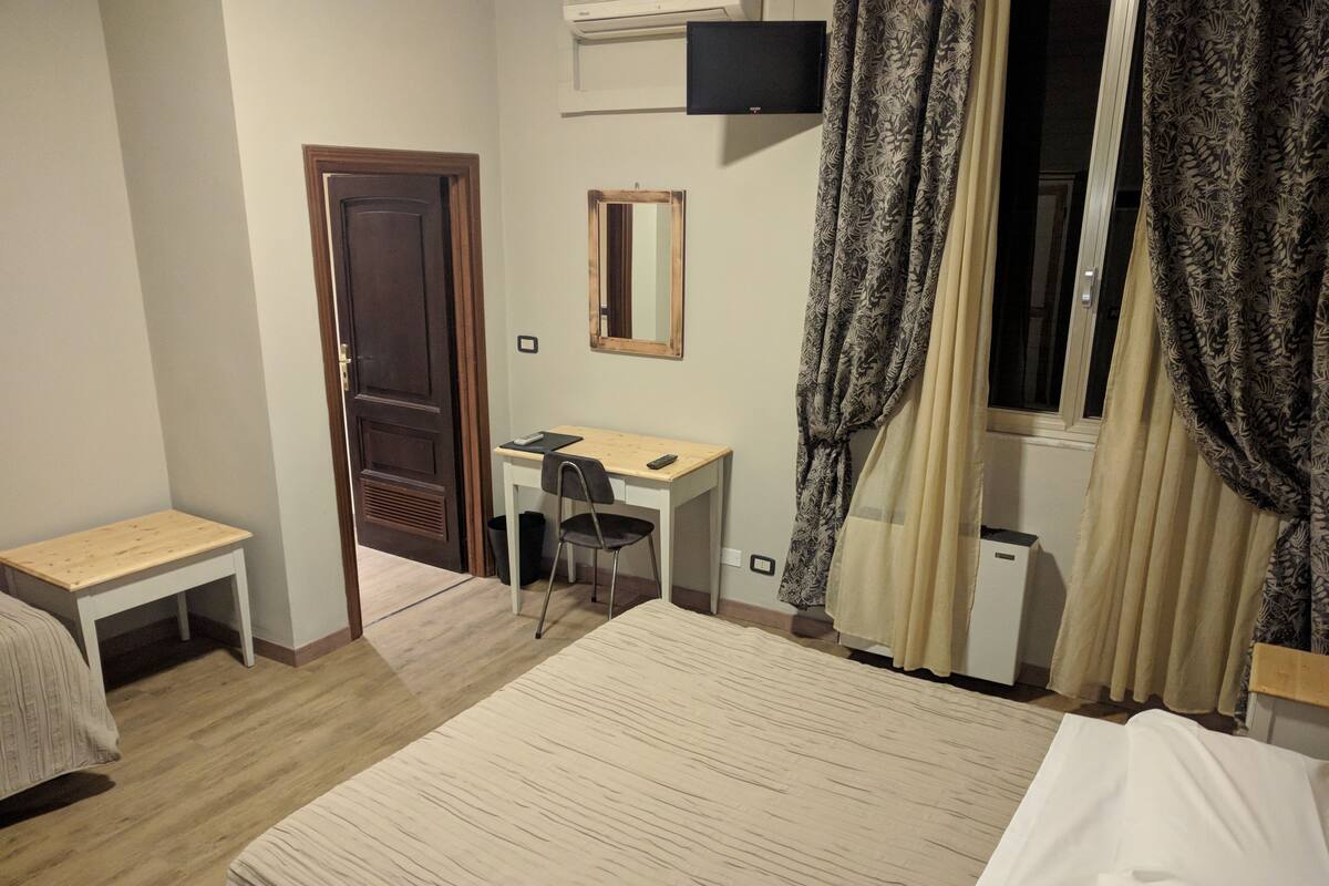 Une chambre d’hôtel comprenant un lit, un bureau, une chaise, une petite table, un miroir et une fenêtre avec des rideaux.
