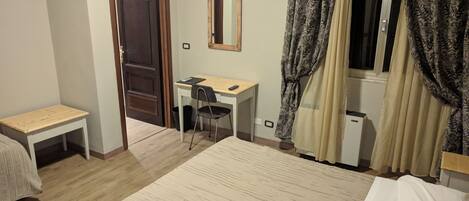 A hotel room with a bed, a desk, a chair, a small table, a mirror, and a window with curtains.