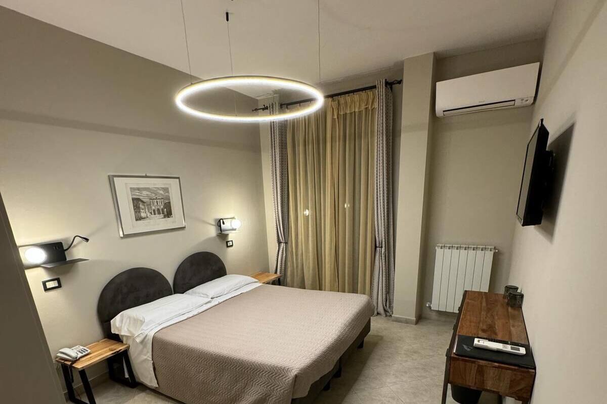 Une chambre d’hôtel moderne équipée d’un lit, de tables de chevet, d’un bureau, d’une chaise, d’un luminaire au plafond, d’un radiateur et d’un tableau encadré au mur.