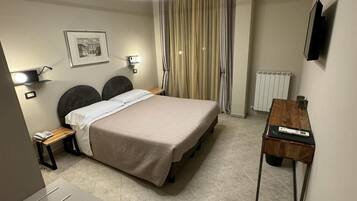 Une chambre d’hôtel moderne équipée d’un lit, de tables de chevet, d’un bureau, d’une chaise, d’un luminaire au plafond, d’un radiateur et d’un tableau encadré au mur.